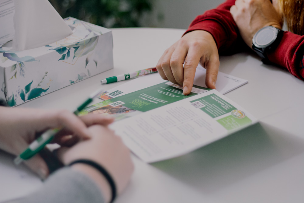 Eine Person im Vordergrund reicht der anderen Person ein Blatt Papier oder eine Karte.